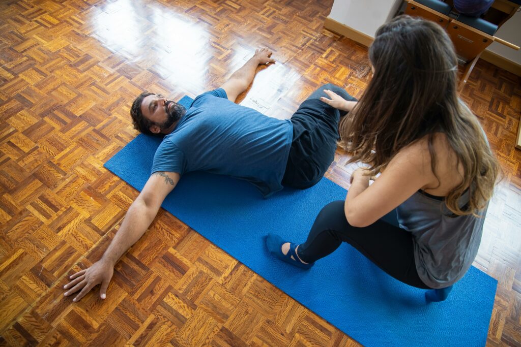 A man and woman engaged in a rehabilitation exercise on a yoga mat indoors.
